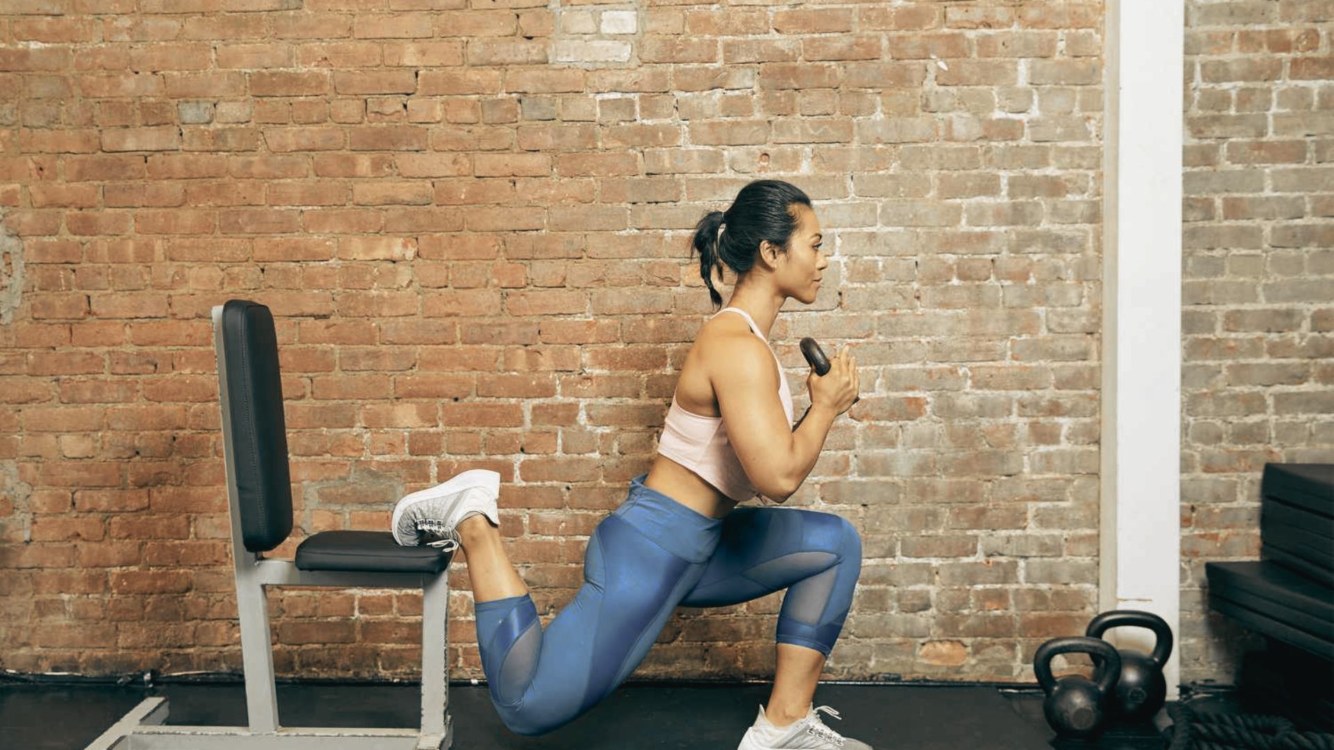 A woman in blue leggings and a sports bra does a Bulgarian split squat with her rear foot on a bench.