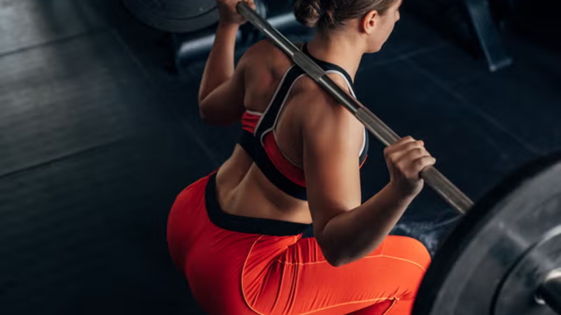 Woman in red leggings and sports bra performing a barbell back squat, viewed from above.