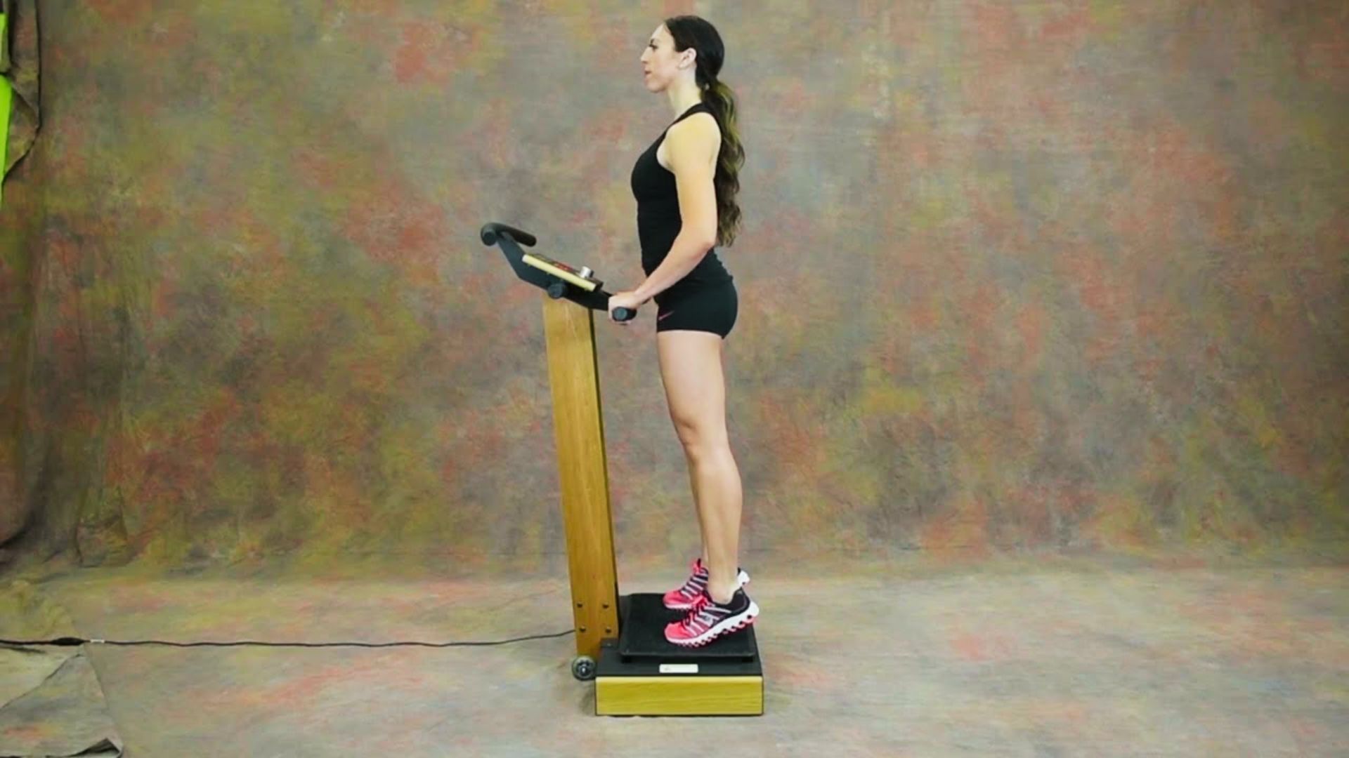 A woman in a black tank top and shorts stands upright on a vibration plate with an attached handlebar in a studio setting.