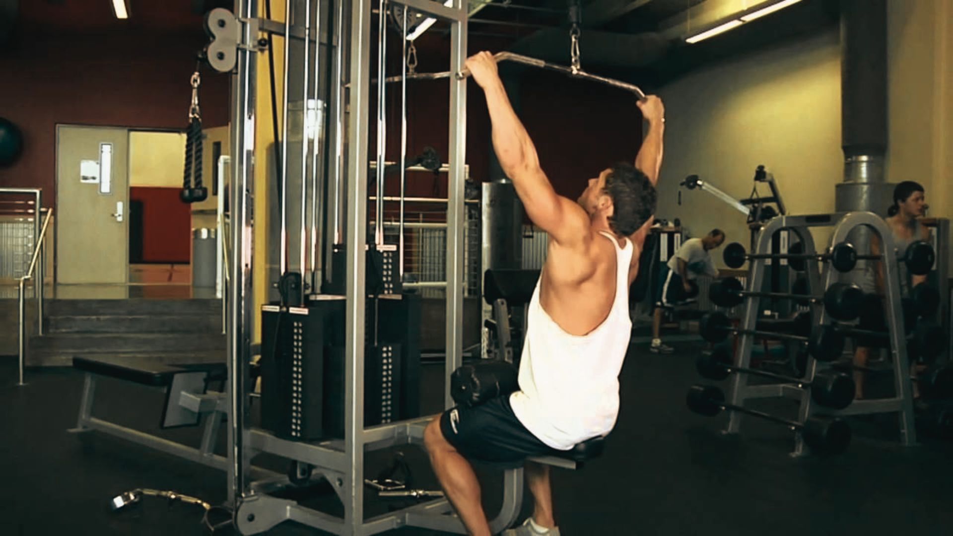Man in a white tank top performing a wide-grip lat pulldown on a cable machine.
