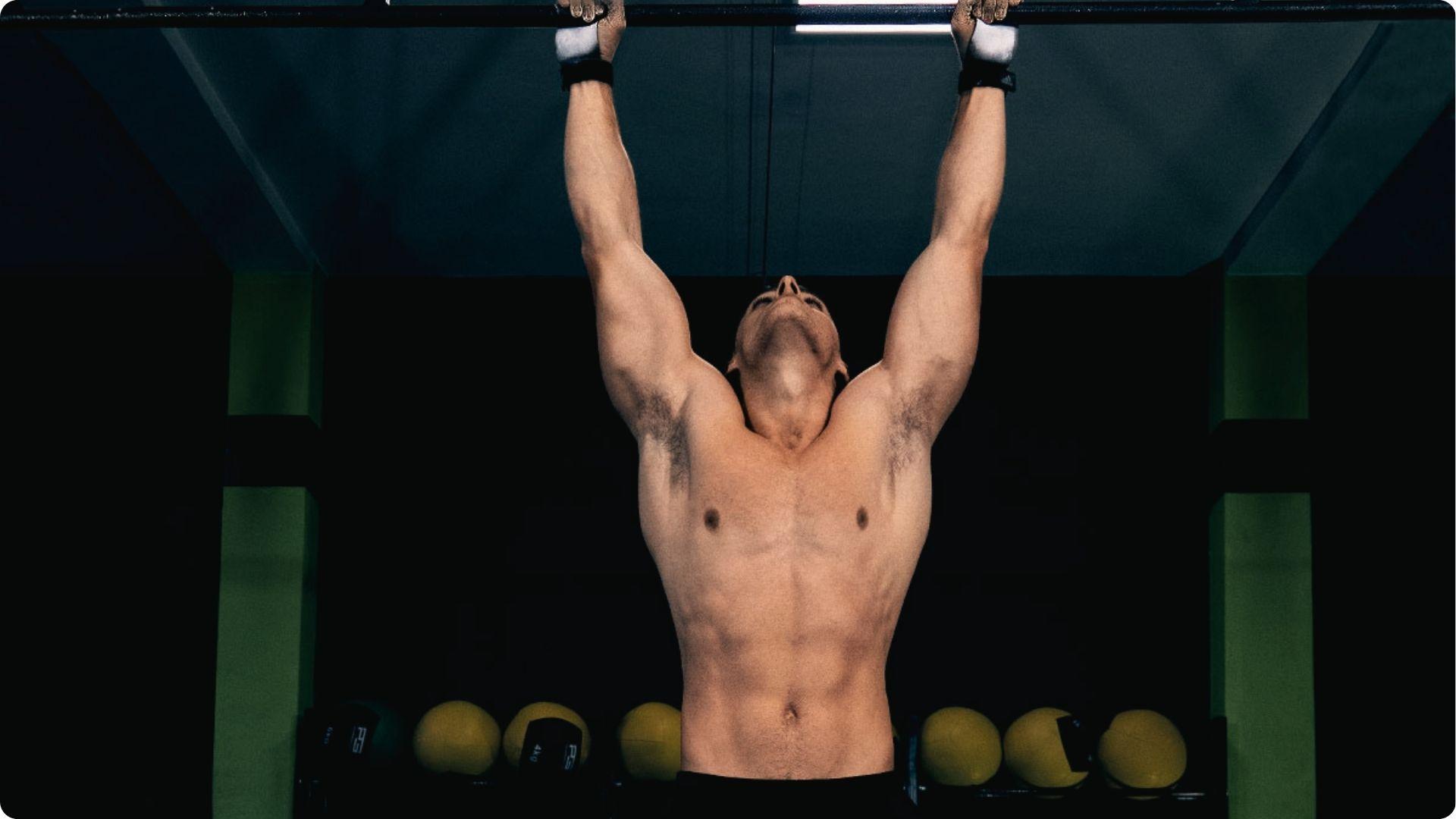 Shirtless man with wrist straps doing pull-up in gym, head tilted back, lats engaged