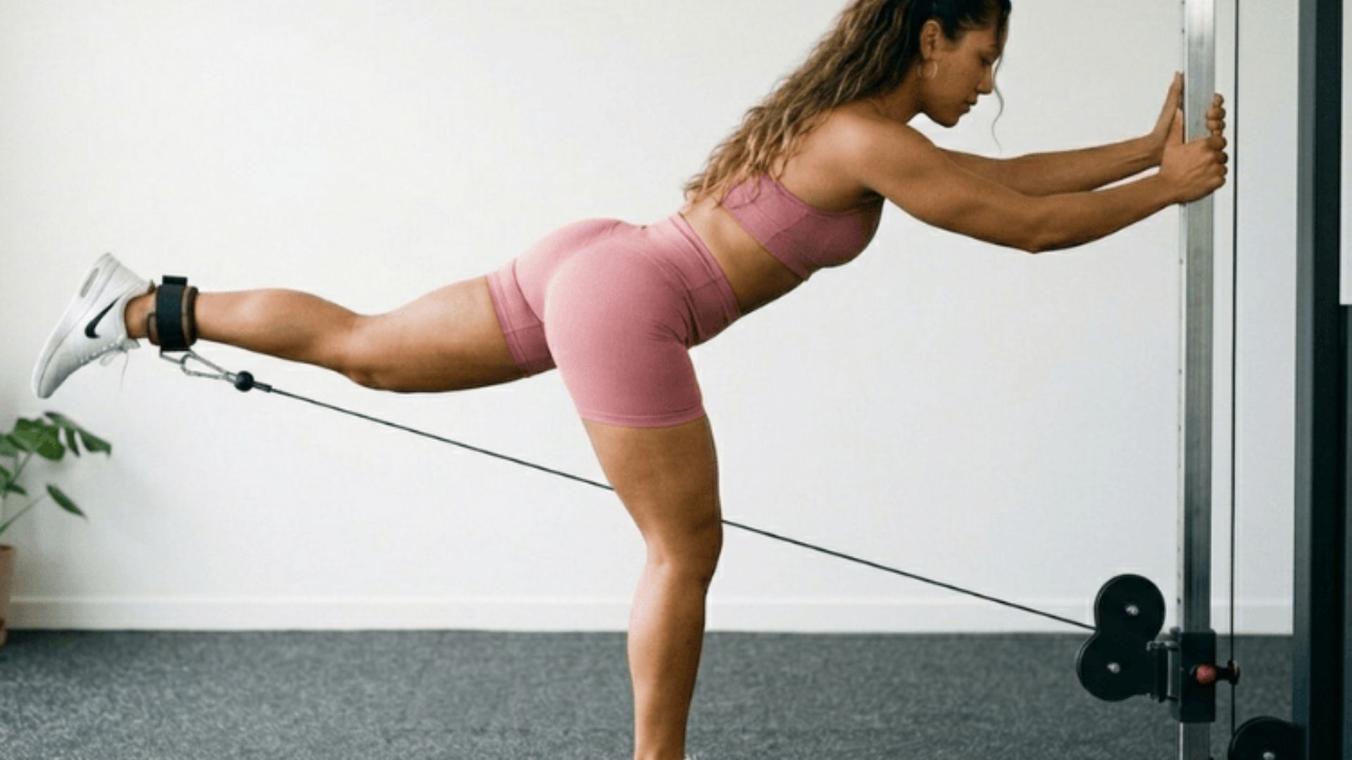 Woman in pink workout set performing a cable glute kickback while holding a pole for stability.