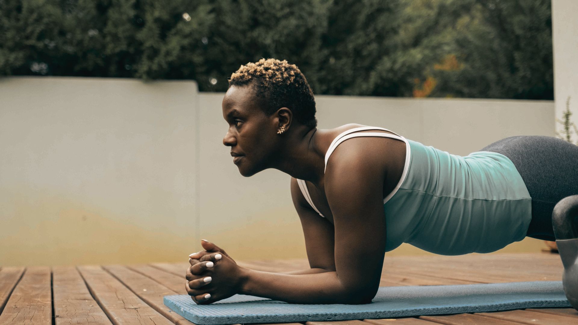 Woman in a teal tank top holds a forearm plank on a blue exercise mat placed on a vibration plate outdoors.