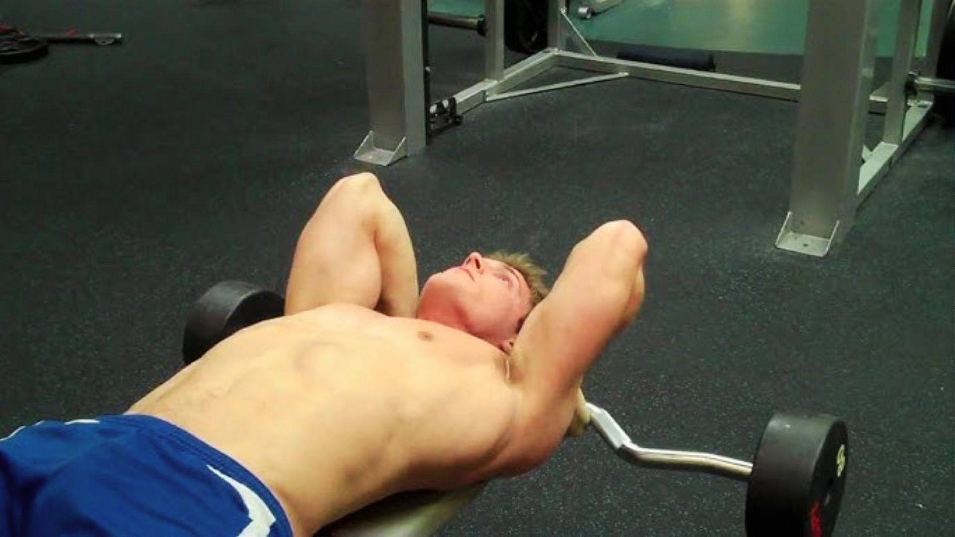 A shirtless man lies on a flat bench doing a barbell lying triceps extension, also called a skull crusher, targeting the triceps.