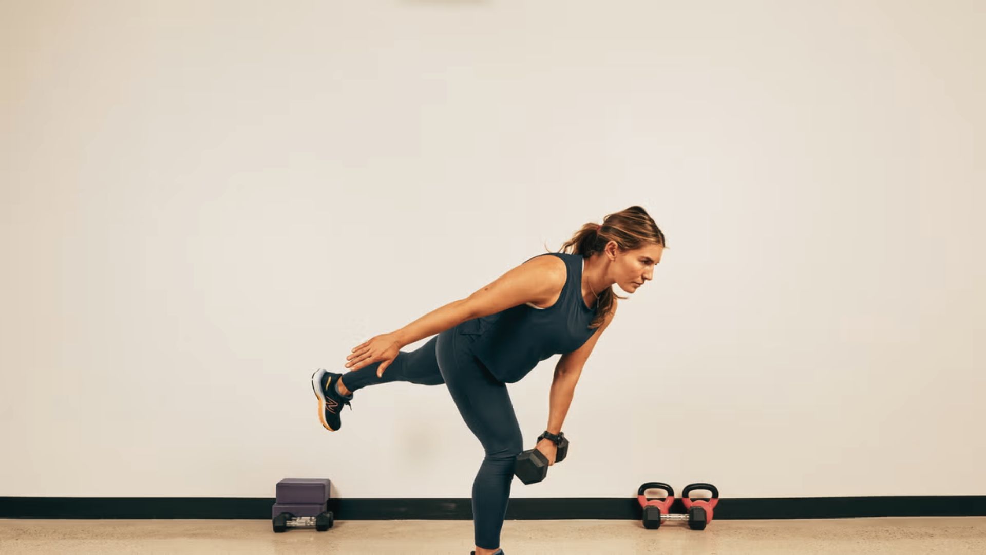 A woman demonstrates a single leg Romanian deadlift in a studio, holding a dumbbell in one hand.