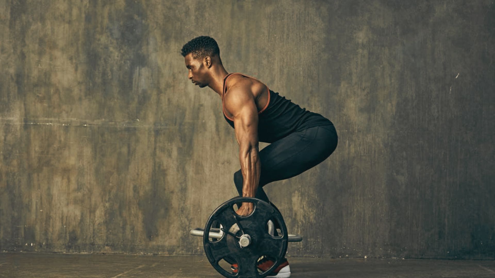Athletic man in a tank top performing a barbell deadlift against a textured wall.