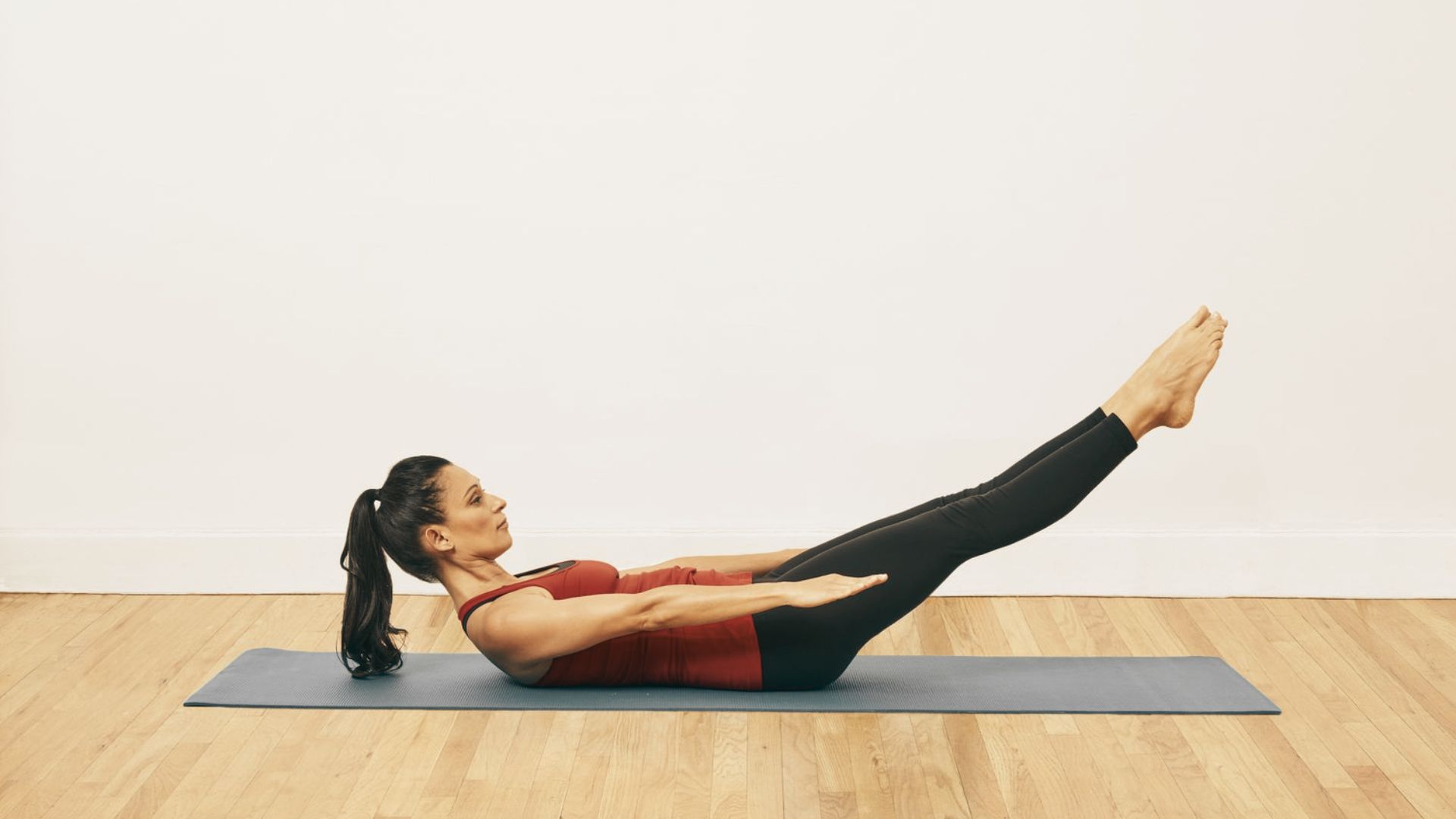 Woman in mauve set on a mat in a Pilates teaser pose with legs extended and arms reaching forward.