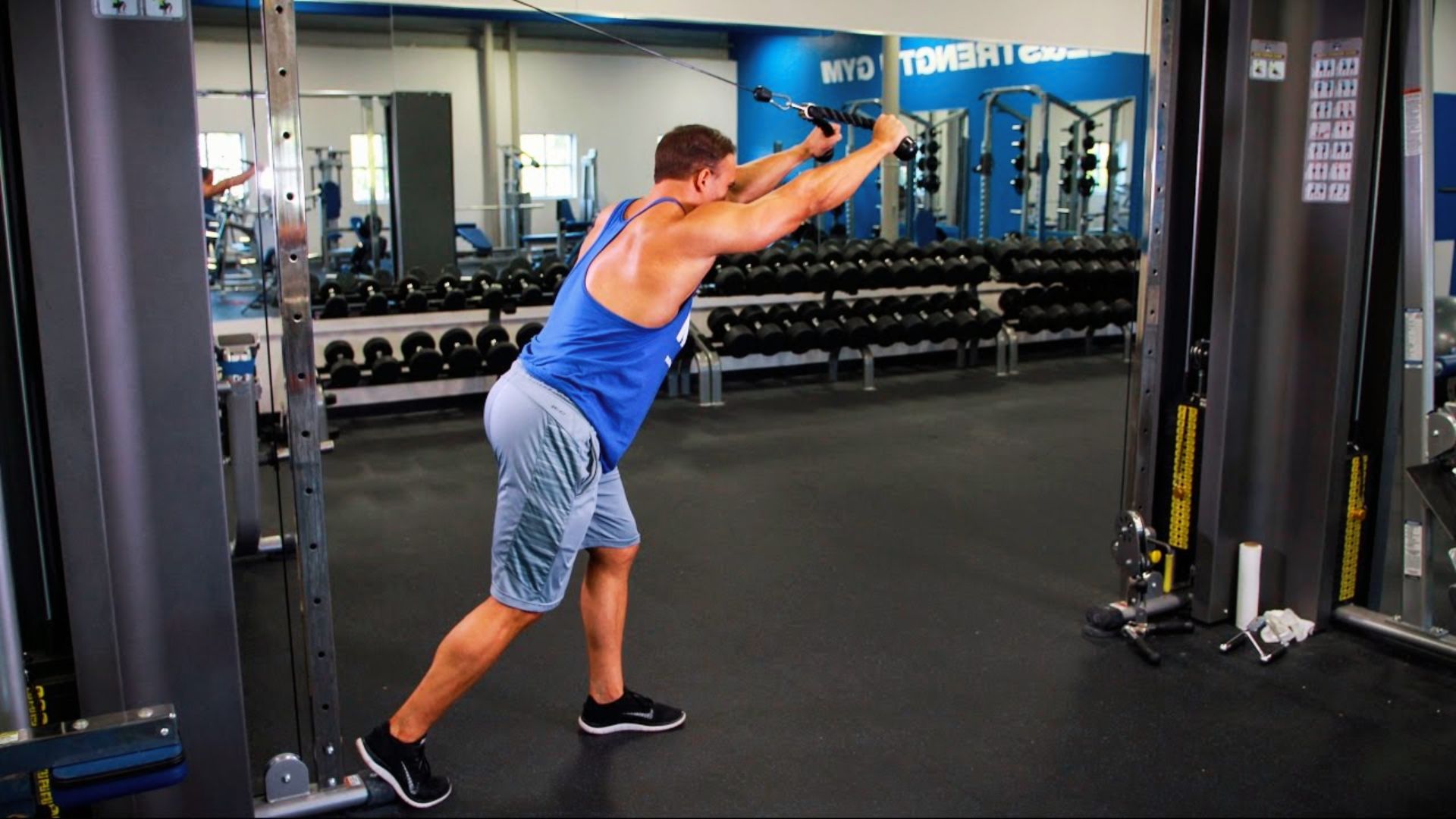 A man in a blue tank top performs a rope cable face pull in a gym, an exercise that engages the rear delts and triceps.