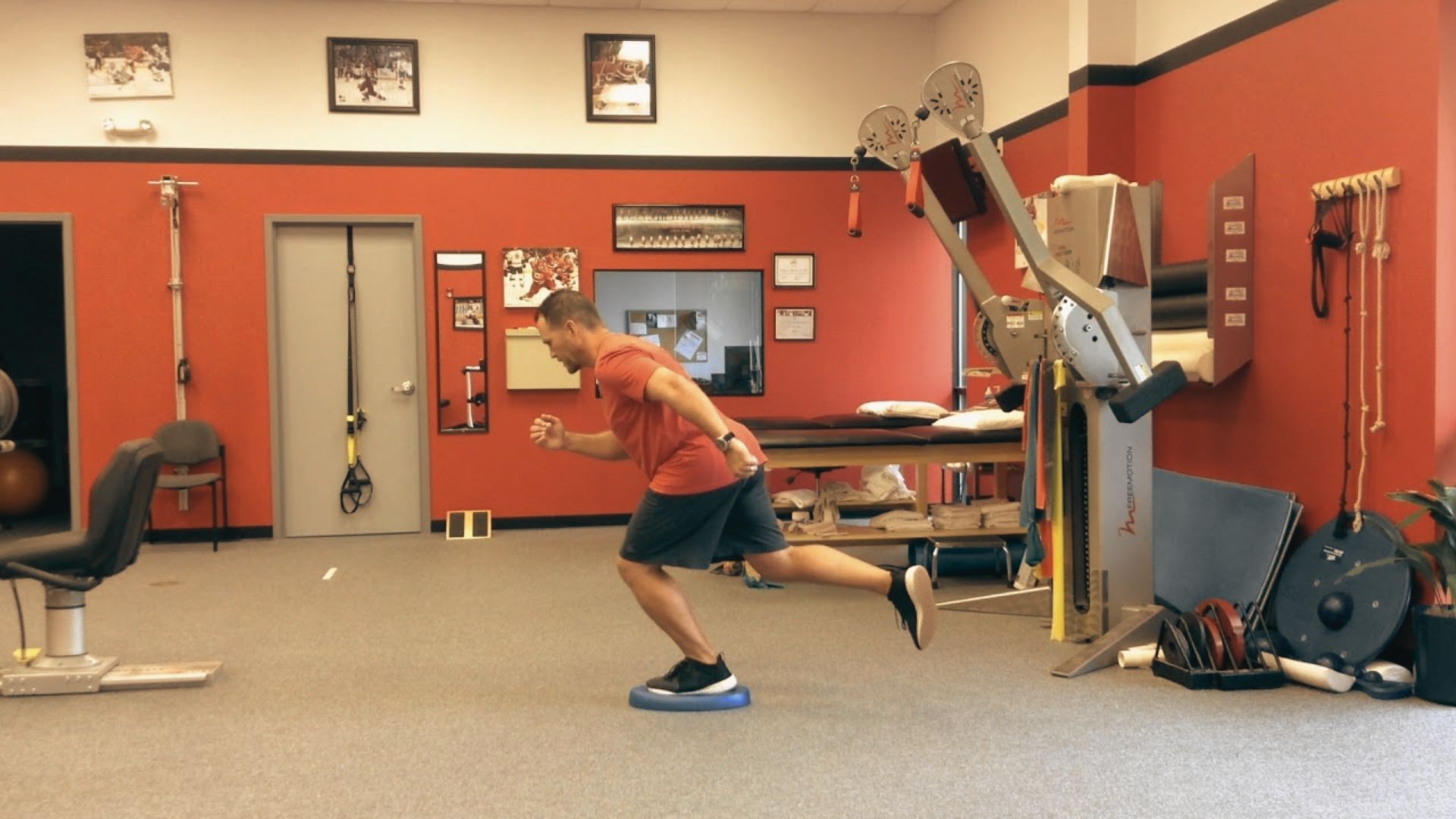 A man in a red shirt balances on one leg atop a blue stability disc in a physical therapy clinic.
