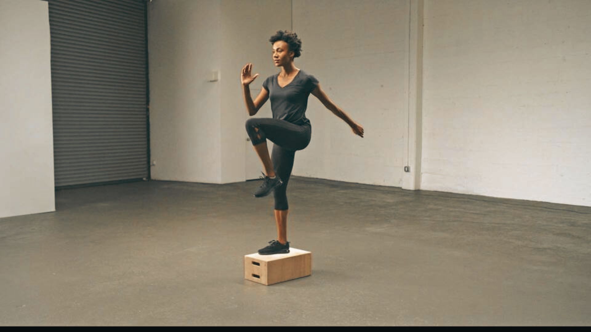 Woman balancing on a wooden plyo box doing a single-leg step-up in a warehouse gym.