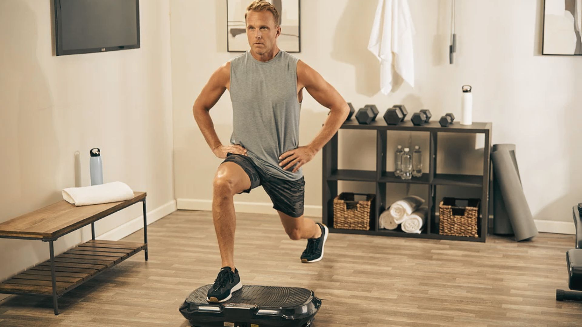 Man in a gray tank top and black shorts does a forward lunge with one foot on a black vibration plate in a home gym.