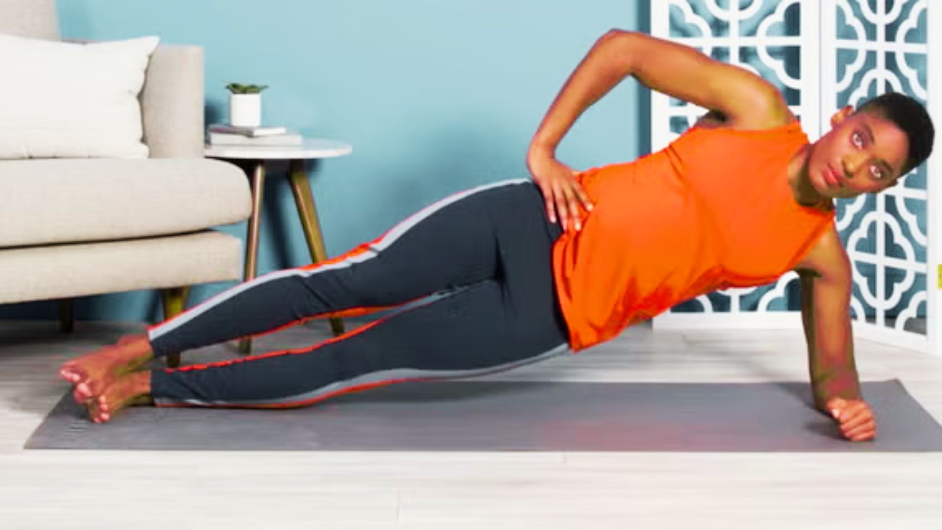 Person in an orange tank top and black leggings performing a Pilates side plank on a gray yoga mat.