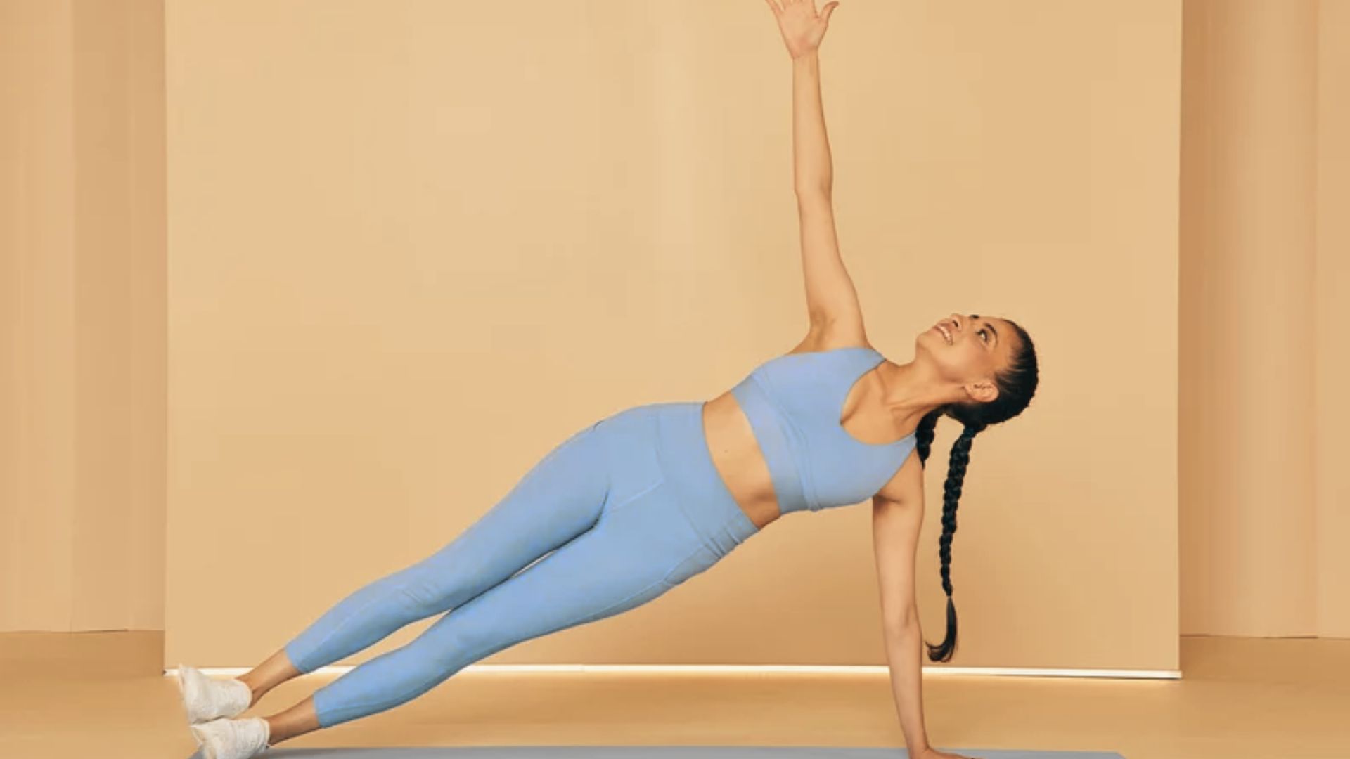 Woman in light blue activewear holds a side plank on a mat, lifting her top arm toward the ceiling against a beige backdrop.