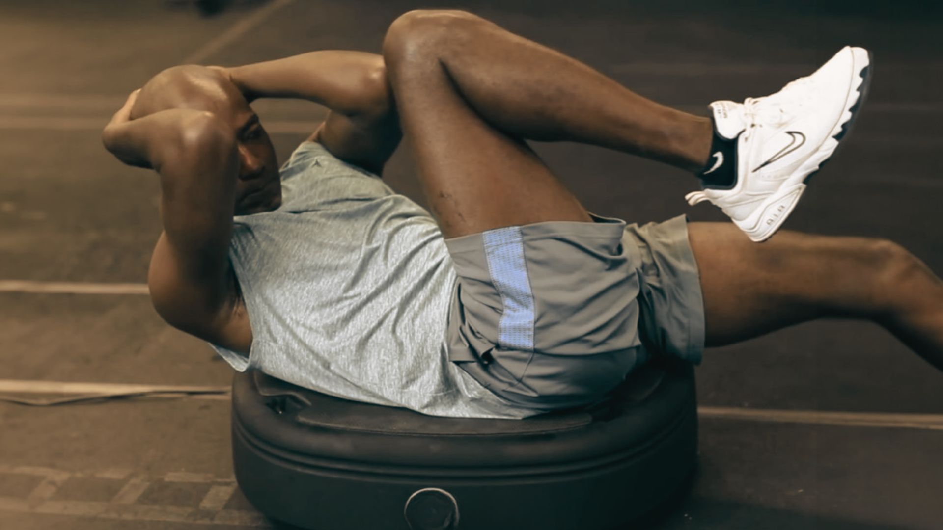 Man in a gray tank top and shorts performs bicycle crunches while seated on a vibration plate.